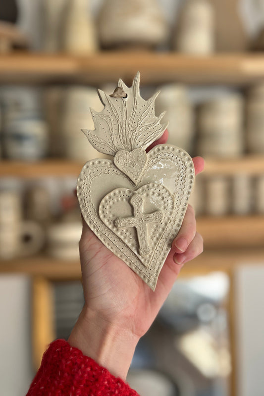 ex voto ou coeur sacré en grès blanc émaillé il est représenté une croix en relief et le coeur est travaillé dans la matière comme martelé. il est tenu dans la main de la céramiste à Montpellier